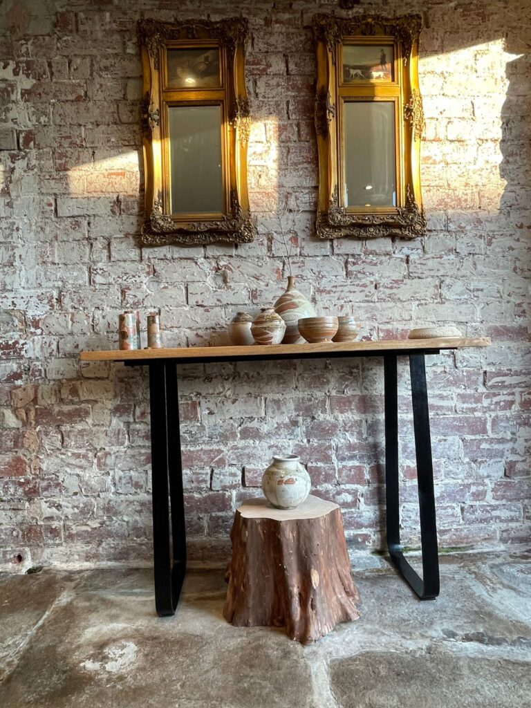 Mirrors, handmade ceramic pottery sat on a side table and a tree trunk with a ceramic pot sat on it, in an entrance hall with exposed stone wall and stone flooring