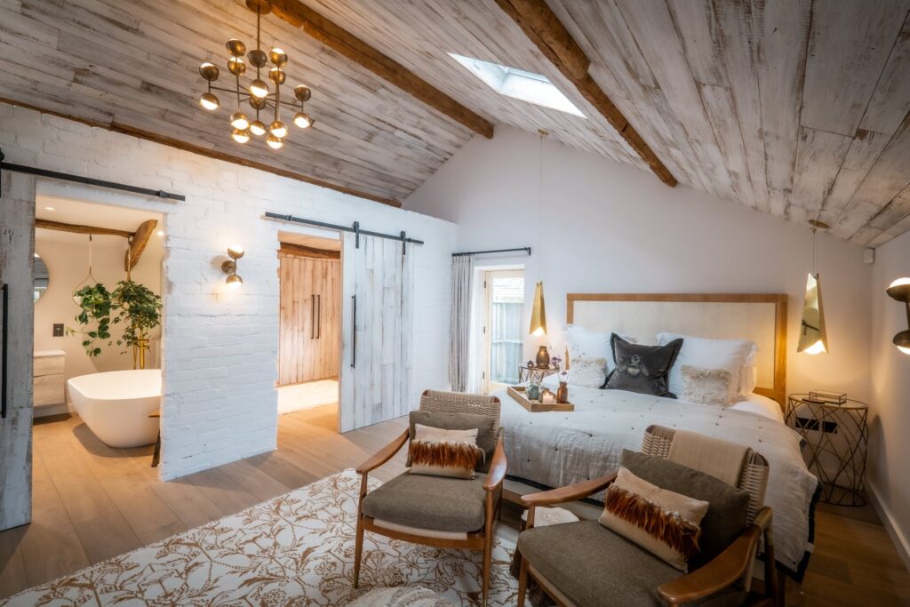 Bedroom with white painted exposed stone walls, ceiling reclaimed wood cladding and wood flooring in a Scandi style with doors into the ensuite and dressing room open