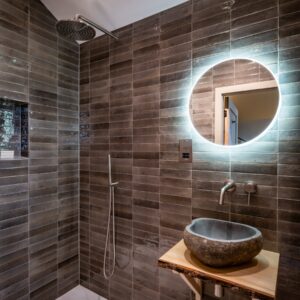 Luxury ensuite bathroom infused with natural materials with a stone pebble sink set on a rustic wooden plinth and a circular illuminated vanity mirror
