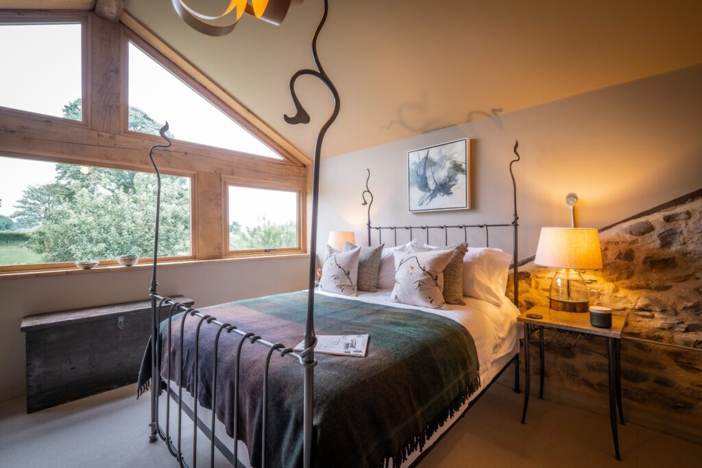 Bedroom design infused with natural materials with an exposed stone wall, wooden beams, an oak frame, bespoke cabinetry and lighting design designed by Charlotte Findlater Design, an award winning interior design studio specialising in luxury, high-end residential & commercial projects that respect time, nature and people.