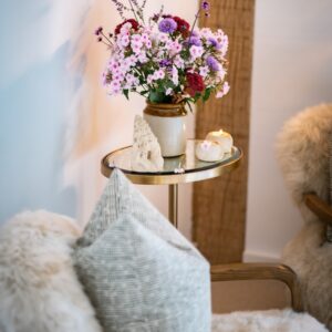 Close up of a case of purple flowers sat on a table next to a comfortable bedroom seat