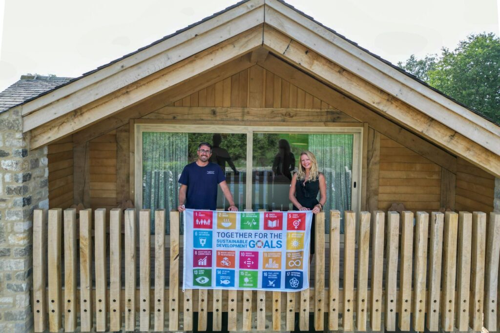 Charlotte Findlater, a Biophilic Interior Designer, and Scott Findlater standing on a balcony holding a large UN 17 Sustainable Development Goals flag
