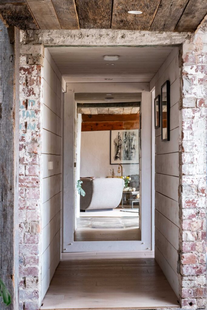 A hallway combining multiple mixed materials, wood floor, exposed red bricks, a concrete lintel, wood panelling and a full height mirror at the end bouncing light around