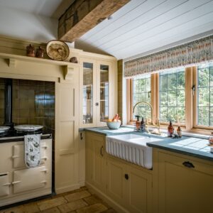 A bespoke farmhouse style kitchen, painted in a custom buttermilk paint colour. There is an Aga, double belfast sink and green slate worktops with a clear view out of the window.