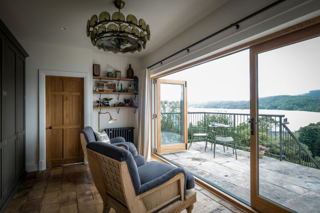 A garden room seating area with terracotta floor tiles, and 2 oversized deconstructed blue cushioned chairs with a view outside to the lake. The bifold doors are wide open.