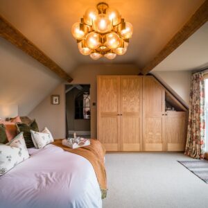 A principle bedroom with a super king bed with a natural wool throw draped, bespoke oak wardrobes, feature glass and metal large sphere cluster pendant and a sliding door to a lake view balcony