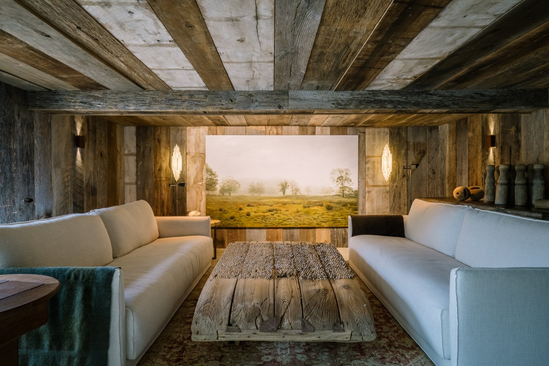 Rustic lounge with two large sofas facing a chunky antique wheat sheaf plough coffee table, landscape artwork and crystal sconces, barn siding clad walls and ceiling