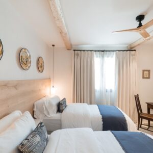 A twin bedroom with marez flooring, with 3 traditional Menorcan hand painted dishes mounted on the wall, hand made wardrobes and an antique desk, mirror and chair in the corner by the windows