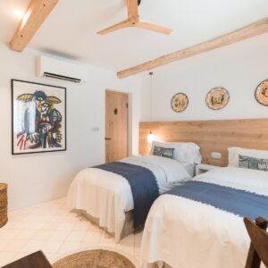 A twin bedroom with marez flooring, with 3 traditional Menorcan hand painted dishes mounted on the wall, wicker baskets and an abstract art piece on the other wall
