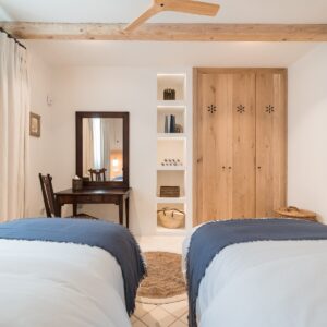 A twin bedroom with hand made wooden wardrobes, abstract wall art and an antique desk, mirror and chair in the corner by the windows