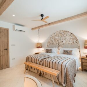 A front on view of the twin bedroom with terracotta and beige bed linen, a recessed arch with a feature stone wall, matching bedside tables and drop pendants, and the hand made wooden bedroom door