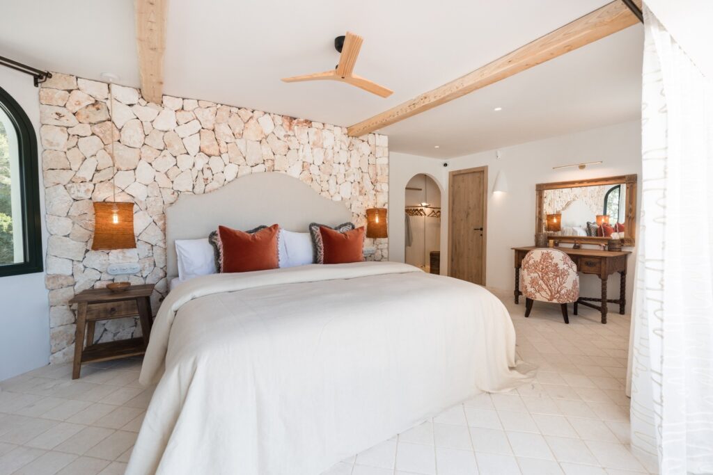 The master ensuite bed set against feature stone wall, with marez flooring, with a glimpse into the dressing room and the antique desk, mirror and chair in the corner