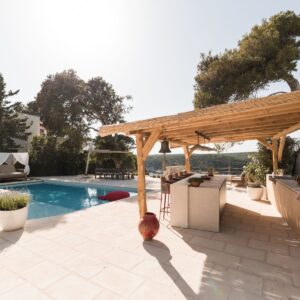 The outdoor terrace of a Menorcan Villa with a pool, wooden framed day bed and large pergola housing a fully furnished outdoor kitchen and bar area