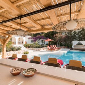 The view from inside a Menorcan villa pergola housing an outdoor kitchen and bar looking across the pool to sun loungers and a daybed