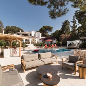 The view across a Menorcan villa terrace, with casual seating for 8 to 10 people, the pool with sun loungers and a daybed, an outside kitchen and bar area, looking back at the villa