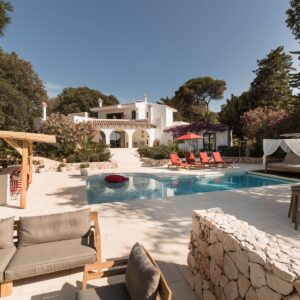 The view across a Menorcan villa terrace, with casual seating for 8 to 10 people, the pool with sun loungers and a daybed, an outside kitchen and bar area, looking back at the villa