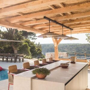 The view from inside a large pergola housing a fully furnished outdoor kitchen and bar area looking into the distant view at a Menorcan villa