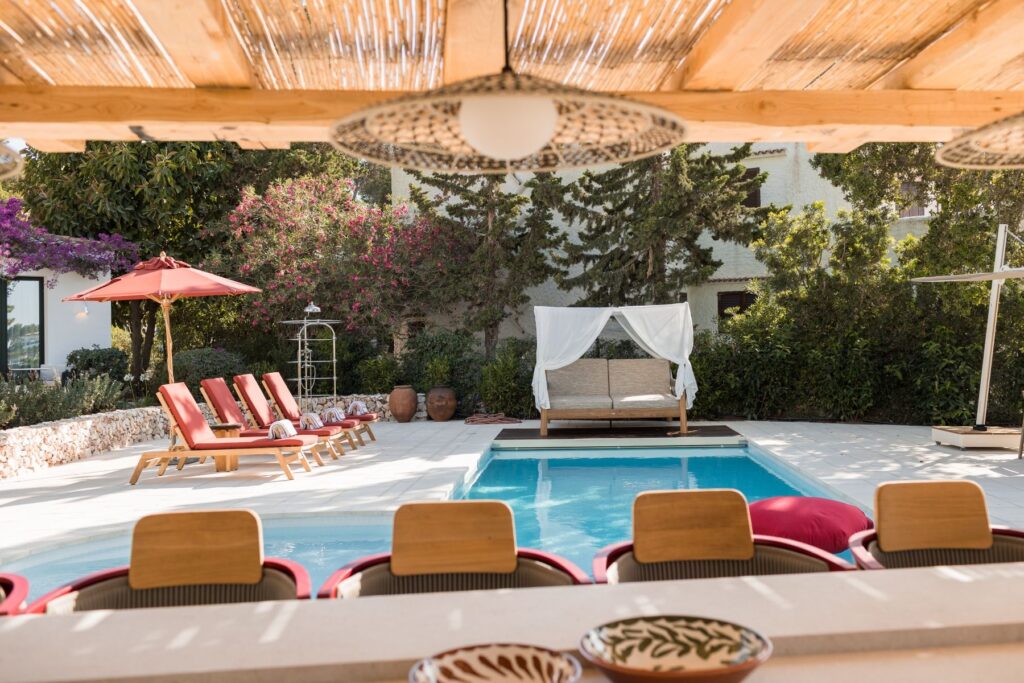 The view from inside a large pergola housing a fully furnished outdoor kitchen and bar area looking across the pool at the daybed and sun loungers at a Menorcan villa