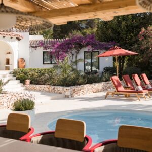 The view from inside a large pergola housing a fully furnished outdoor kitchen and bar area looking across the pool at the sun loungers and the Menorcan villa