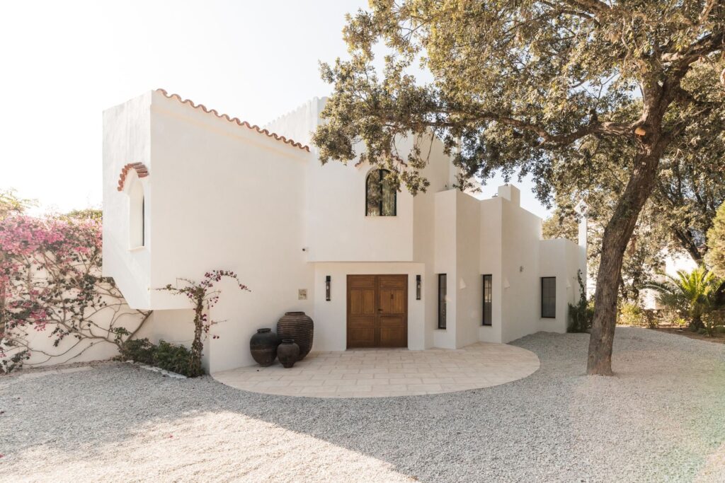 A wide angle view of a crisp white Menorcan villa, simple gravel driveway, with large pots outside the handmade entrance doors