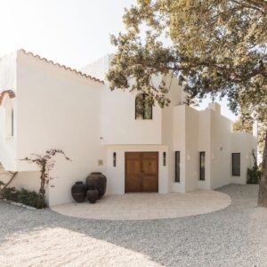 A wide angle view of a crisp white Menorcan villa, simple gravel driveway, with large pots outside the handmade entrance doors