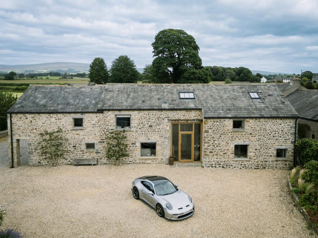 Aerial view of a restored stone barn with slate roof and oak-framed entrance, gravel courtyard, and a silver 911 GT3 Touring parked in front