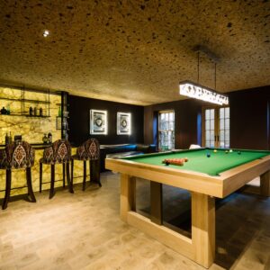 A luxury games room with natural wood flooring, cork ceiling, a custom pool table and a back lit stone wall bar with bar stools.
