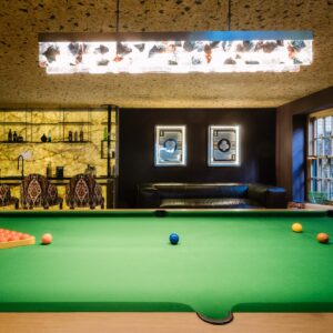 A luxury games room view across the custom pool table to the large wall mounted metal car art pieces and a custom back lit stone wall bar.