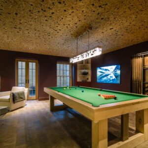A luxury games room with natural wood flooring, cork ceiling, a custom pool table, a large wall mounted TV and custom cue & ball wall mounted cabinetry.