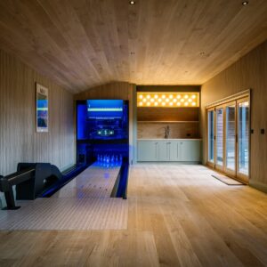 Games room with a bespoke duck pin bowling lane, timber clad ceiling, striped wall finish, glazed doors, and a bar area with sink and illuminated 3D-printed duck pin-shaped lights.