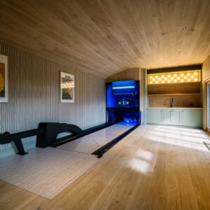 Games room with a bespoke duck pin bowling lane, timber clad ceiling, striped wall finish with framed prints, glazed doors, and a bar area with sink and illuminated 3D-printed duck pin-shaped lights.