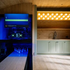 Games room with a bespoke duck pin bowling lane and digital scoring screen, timber clad ceiling, striped wall finish, and a bar area with shaker cabinetry and sink, lit by illuminated 3D-printed duck pin-shaped lights.