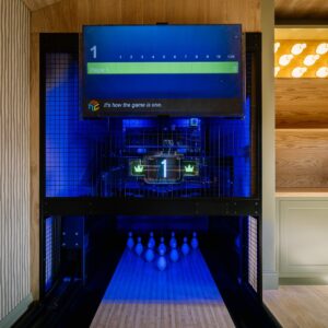 Games room close-up of a bespoke duck pin bowling lane with digital scoring screen and illuminated pins, beside a bar area with shaker cabinetry, sink, and a feature wall of illuminated 3D-printed duck pin-shaped lights.