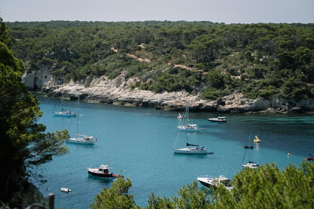 Menorca coastline with turquoise water, sailing boats, rocky cliffs and pine forest, reflecting the island landscape that shapes the design approach.