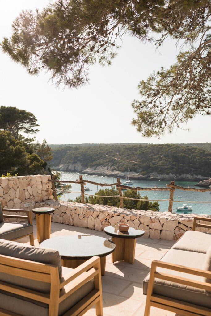 Property renovation in Menorca terrace with natural stone walling, timber furniture and views across a sheltered bay surrounded by pine trees.