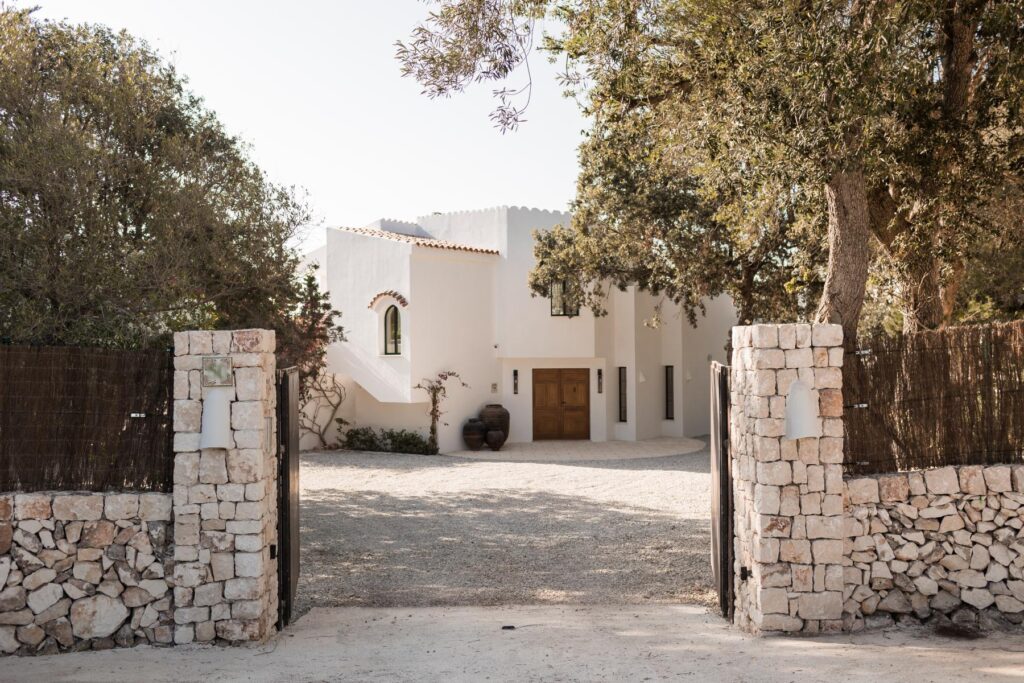 Property renovation in Menorca with a whitewashed entrance, dry stone boundary walls, timber doors and mature trees framing the approach.